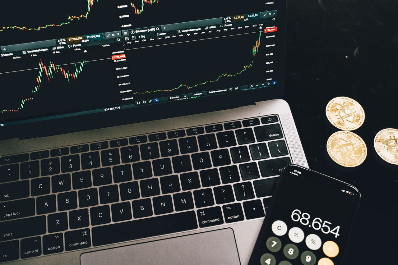 about-us-02 A laptop with trading charts, smartphone calculator, and bitcoin coins depicting cryptocurrency trading.